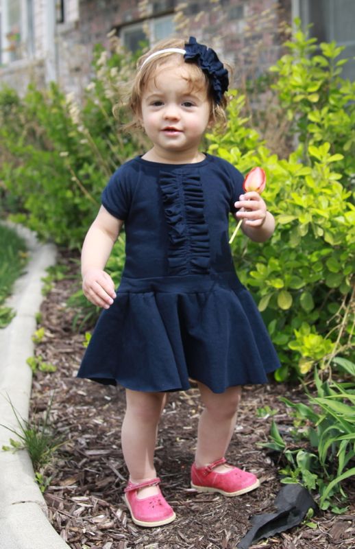 A little girl standing in a garden wearing a blue dress with a ruffle down the front and a circle skirt
