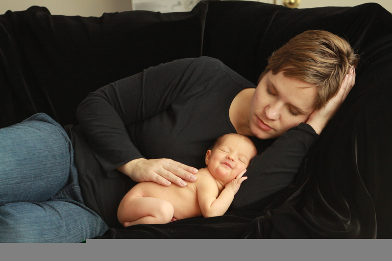 photo of a mom in a black long sleeve t-shirt lying on a couch that\'s covered in black fabric with her baby