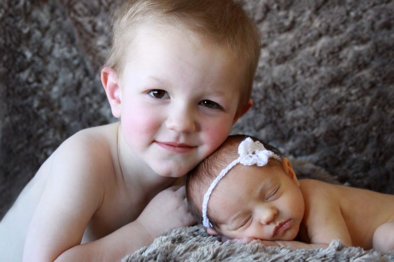 A toddler boy leaning close to his baby sister for a photo