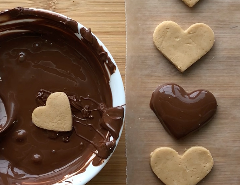 Peanut butter hearts on wax paper with melted chocolate, one heart dipped in chocolate
