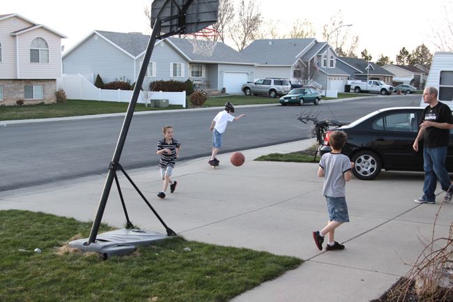 A group of boys in a driveway playing basketball with houses and cars in the background