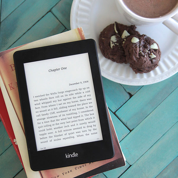 A kindle on a stack of books on a tray with cookies and cocoa