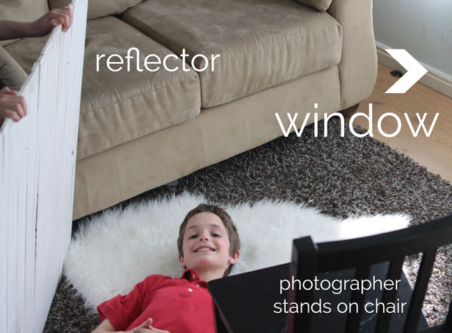 little boy laying on a rug, with white reflector board next to him, and chair for photographer to stand on