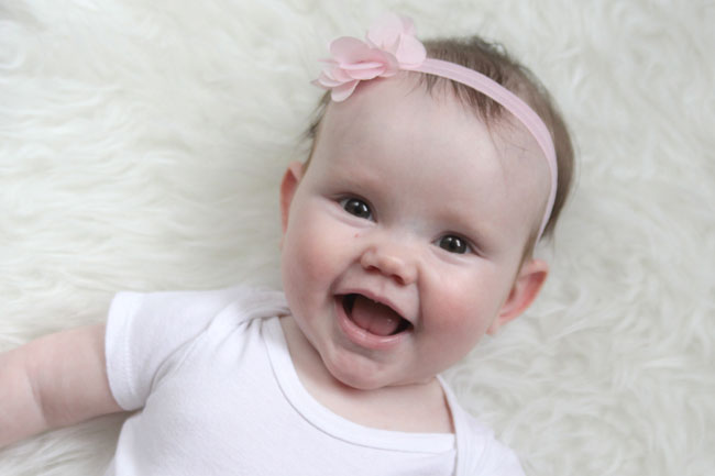 A baby girl laying on a white fur rug smiling at the camera