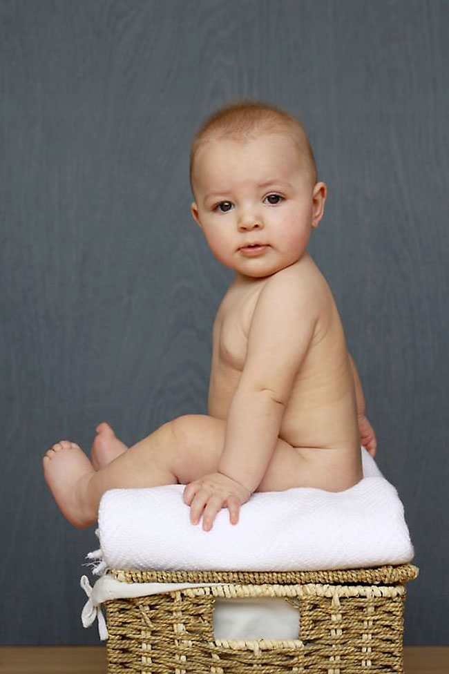 A baby sitting on a wicker basket covered with a blanket