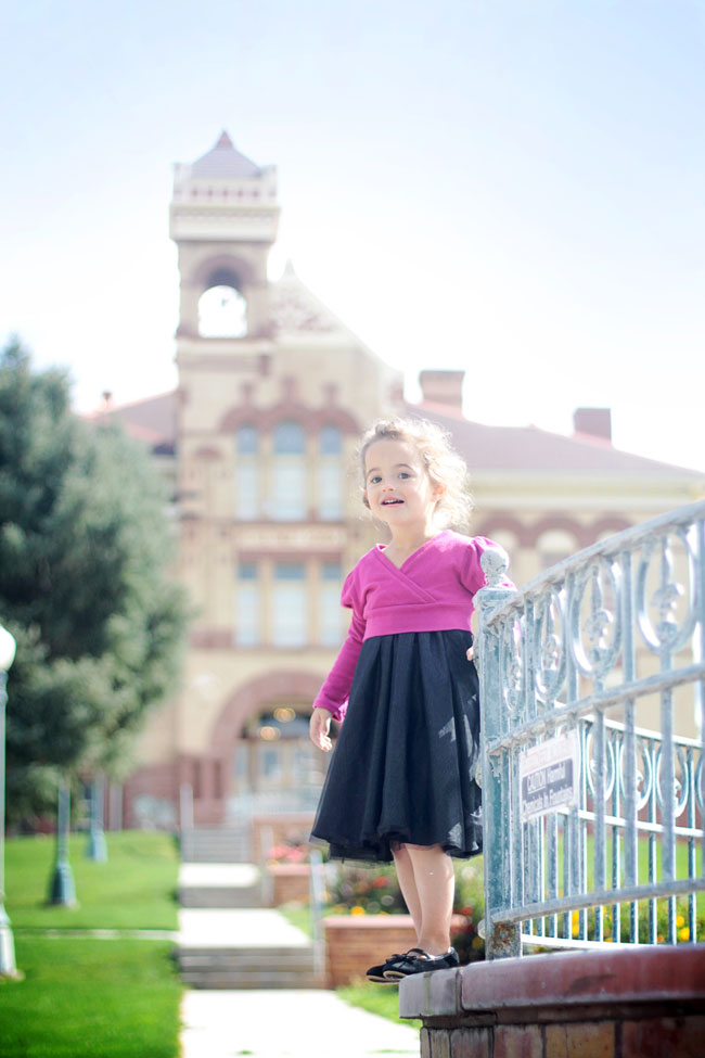 A little girl standing in front of a large building wearing a black dress and pink sweater