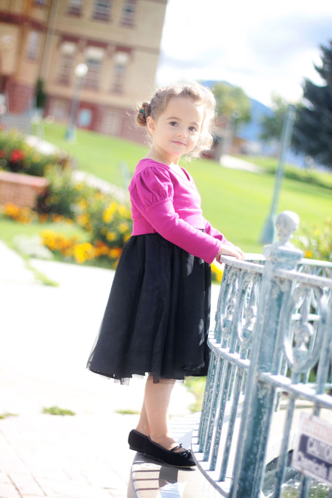 A little girl standing in front of a fence with black skirt and pink sweater