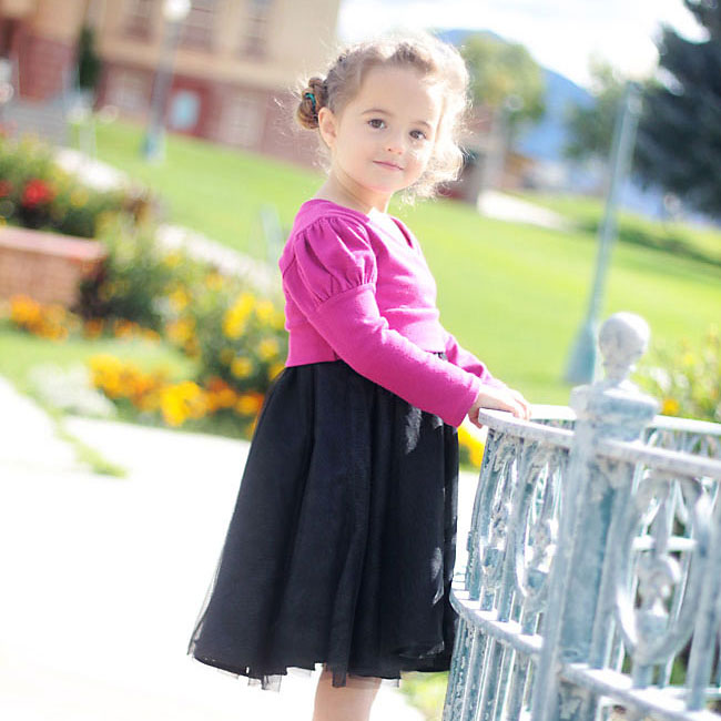 A little girl standing next to a fence wearing a black dress and pink sweater