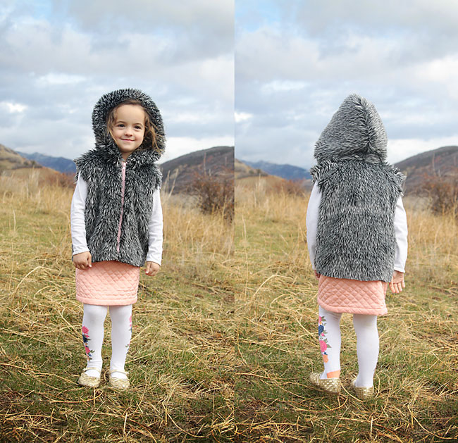 A little girl standing in a field wearing a faux fur vest with a hood