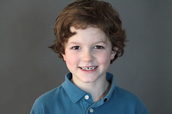 A young boy wearing a blue shirt and smiling at the camera with grey background paper