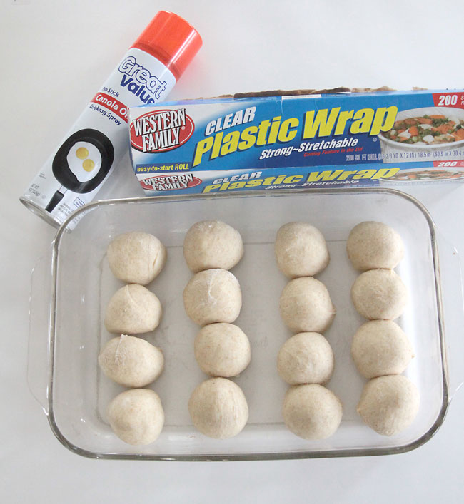 ball of roll dough in a 9x13 glass pan with cooking spray and plastic wrap