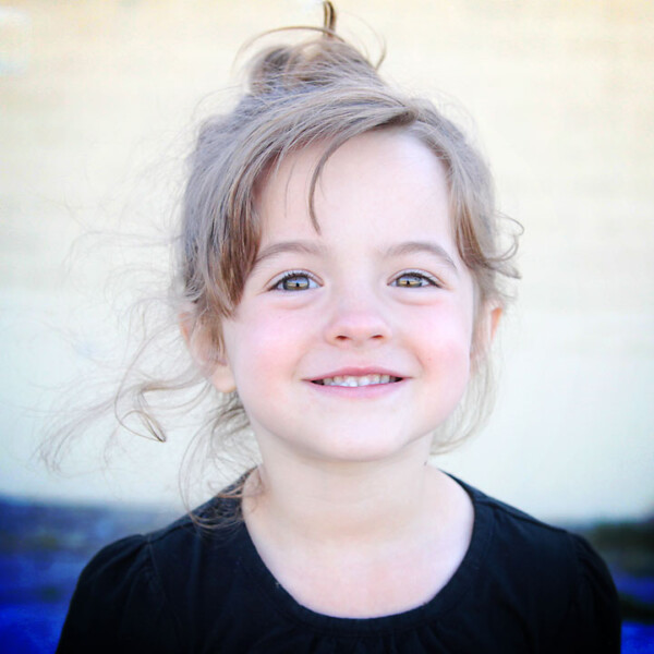 Little girl smiling at the camera in front of a brick wall that's blurred in the background