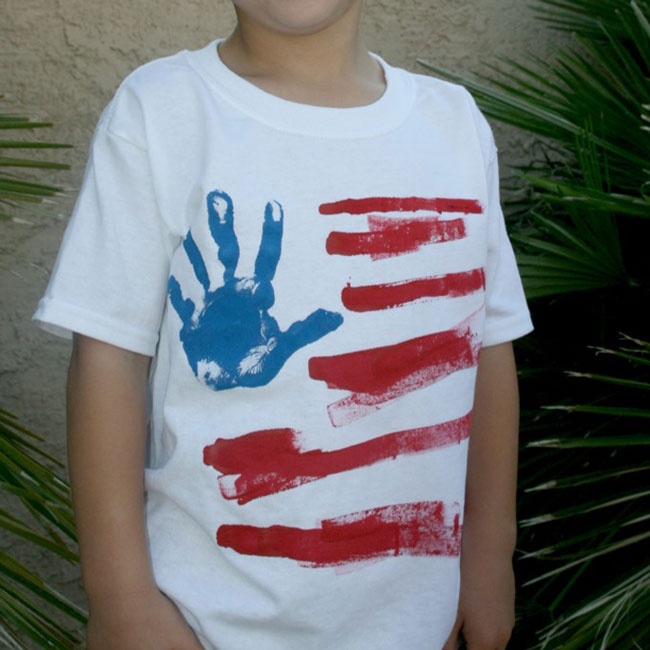A boy wearing a white t-shirt decorated to look like a flag with a blue handprint and red strips for 4th of July