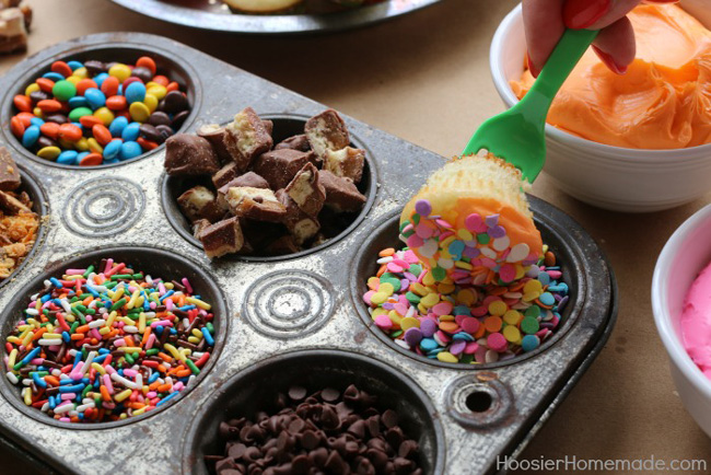 A muffin tin filled with different kinds of candy; a fork dipping a frosted mini cupcake into the candy