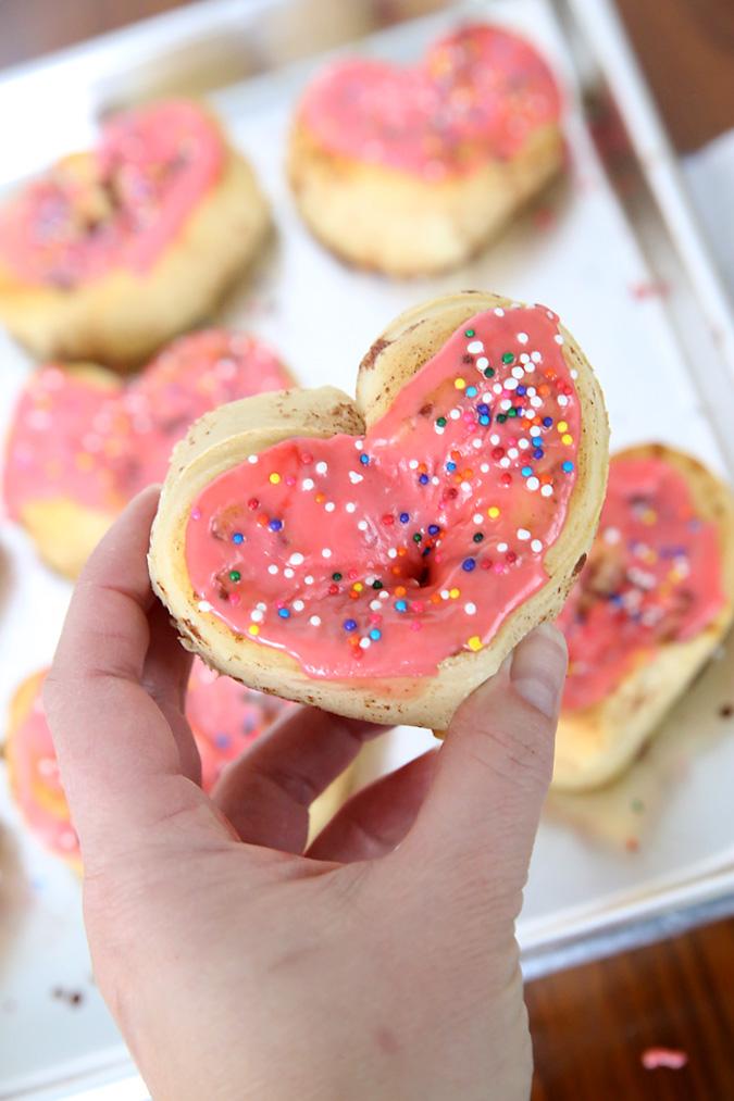 Hand holding a heart shaped cinnamon roll with pink frosting