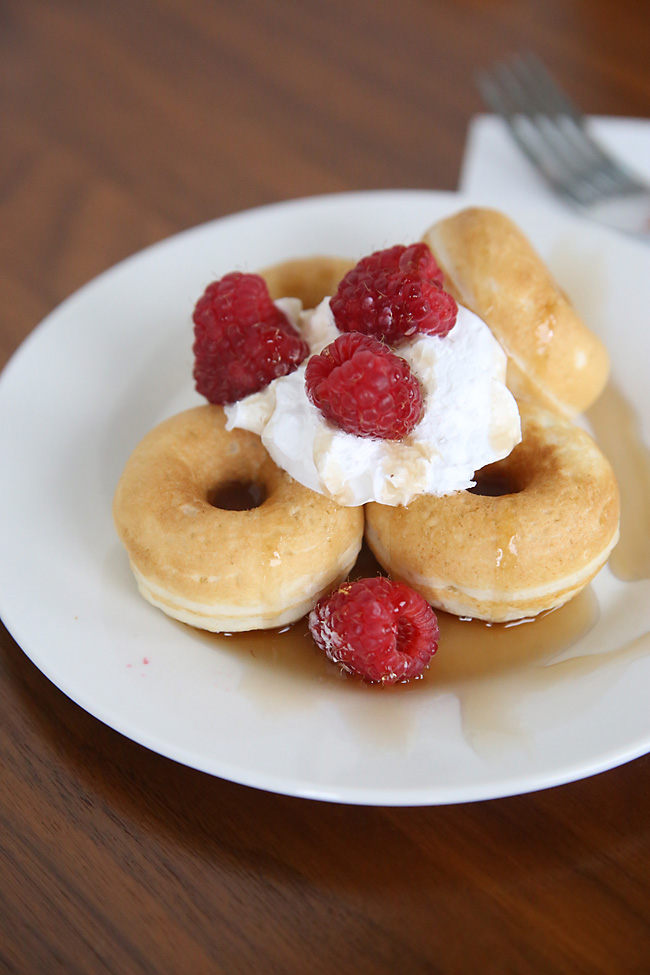 Pancake donuts stacked up on a plate with syrup, whipped cream and raspberries