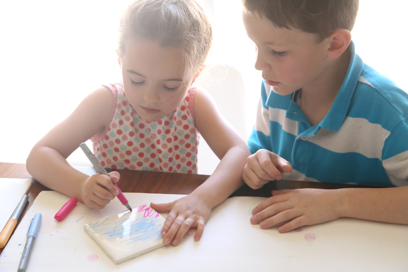 Two children coloring on a white tile to make fired tile art