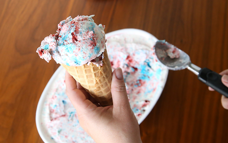 Hand holding a cone with vanilla ice cream with red and blue cake chunks in it
