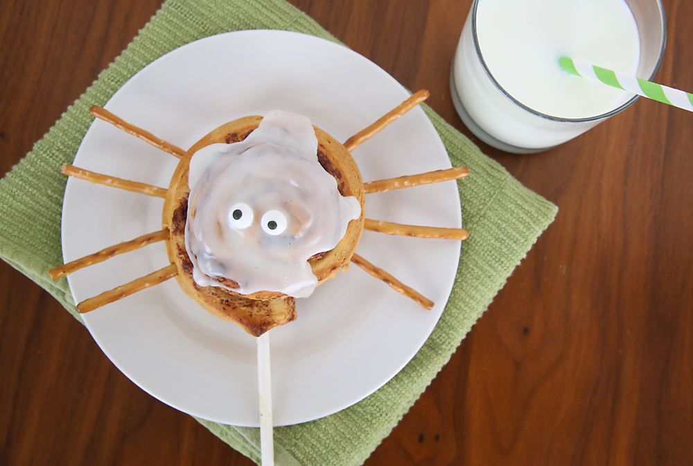 A cinnamon roll on a stick decorated with eyes and pretzel stick legs to look like a spider