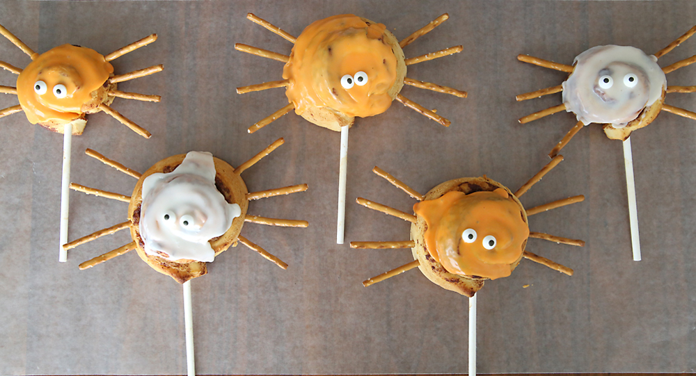A cinnamon roll on a stick decorated with eyes and pretzel stick legs to look like a spider with orange frosting