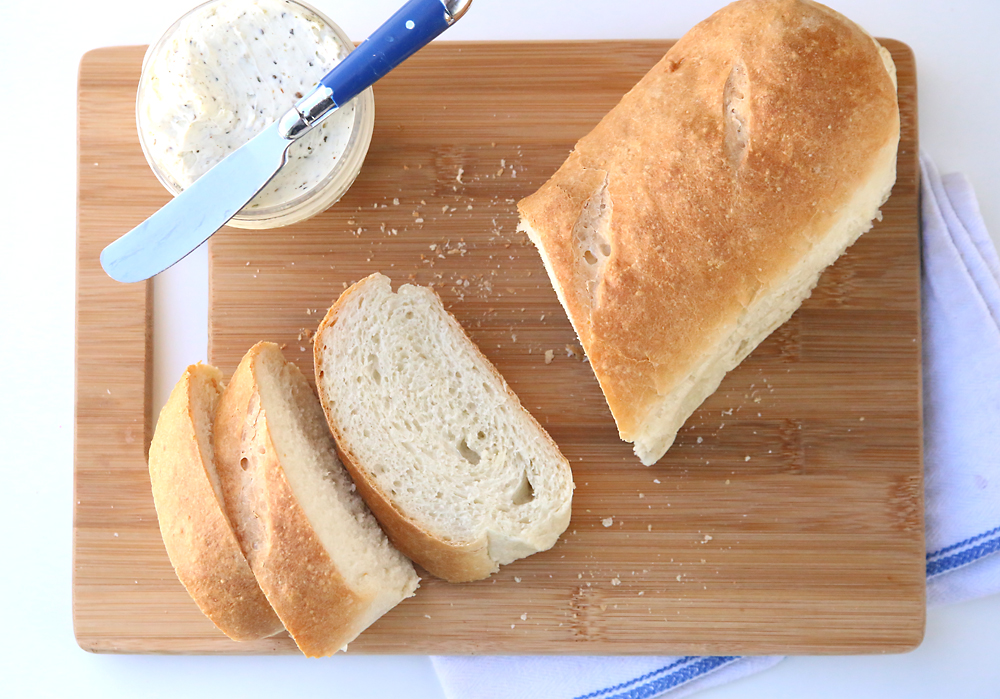 A loaf of french bread slices on a cutting board with a knife and garlic butter in a jar