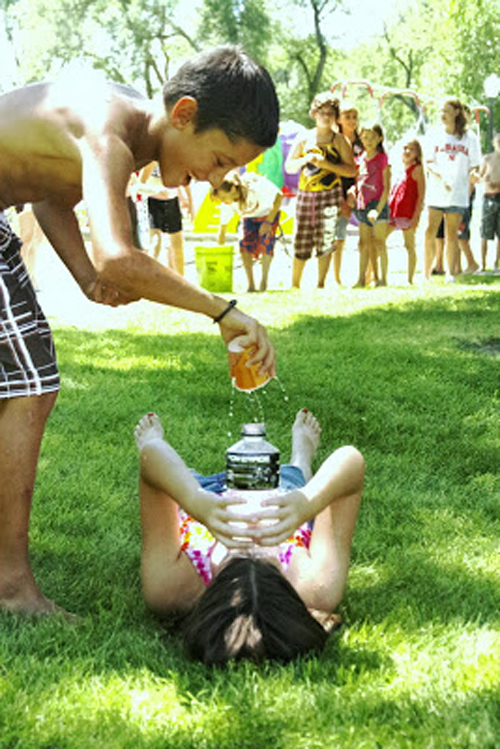 Water games: A boy pouring a cup of water on a girl lying on the grass