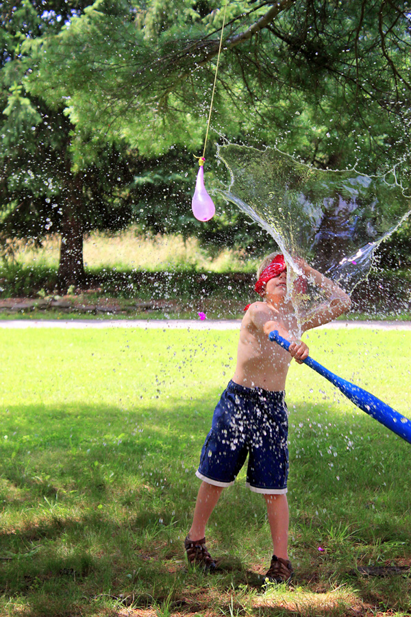 Water games: A young boy using a bat to pop a water balloon