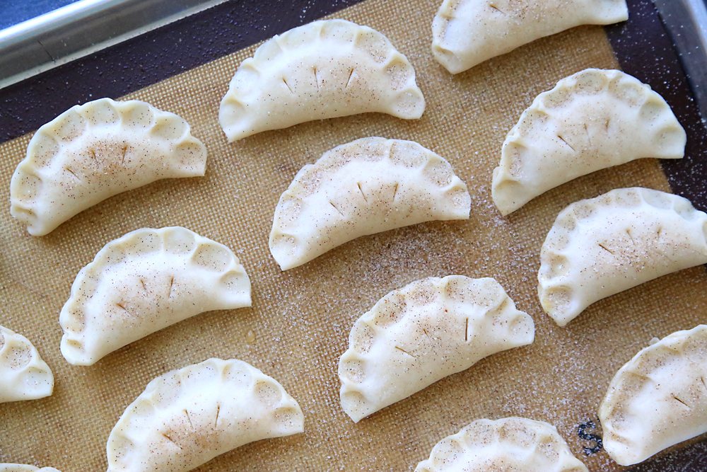 Circles of pie crust filled with apple pie filling and folded over and crimped, on a baking sheet