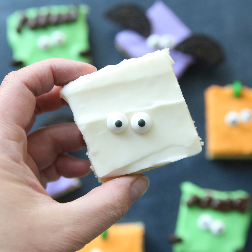 Hand holding a sugar cookie bar with white frosting and two candy eyes