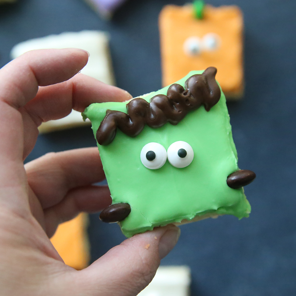 Hand holding a sugar cookie bar with green frosting decorated to look like Frankenstein: chocolate across the top for hair, two candy eyes, and two brown M&Ms for neck bolts