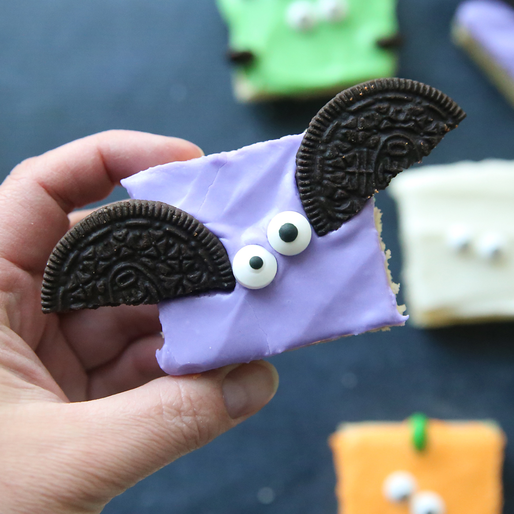 Hand holding a sugar cookie bar with purple frosting, two candy eyes, and an Oreo cookie cut in half to look like bat wings