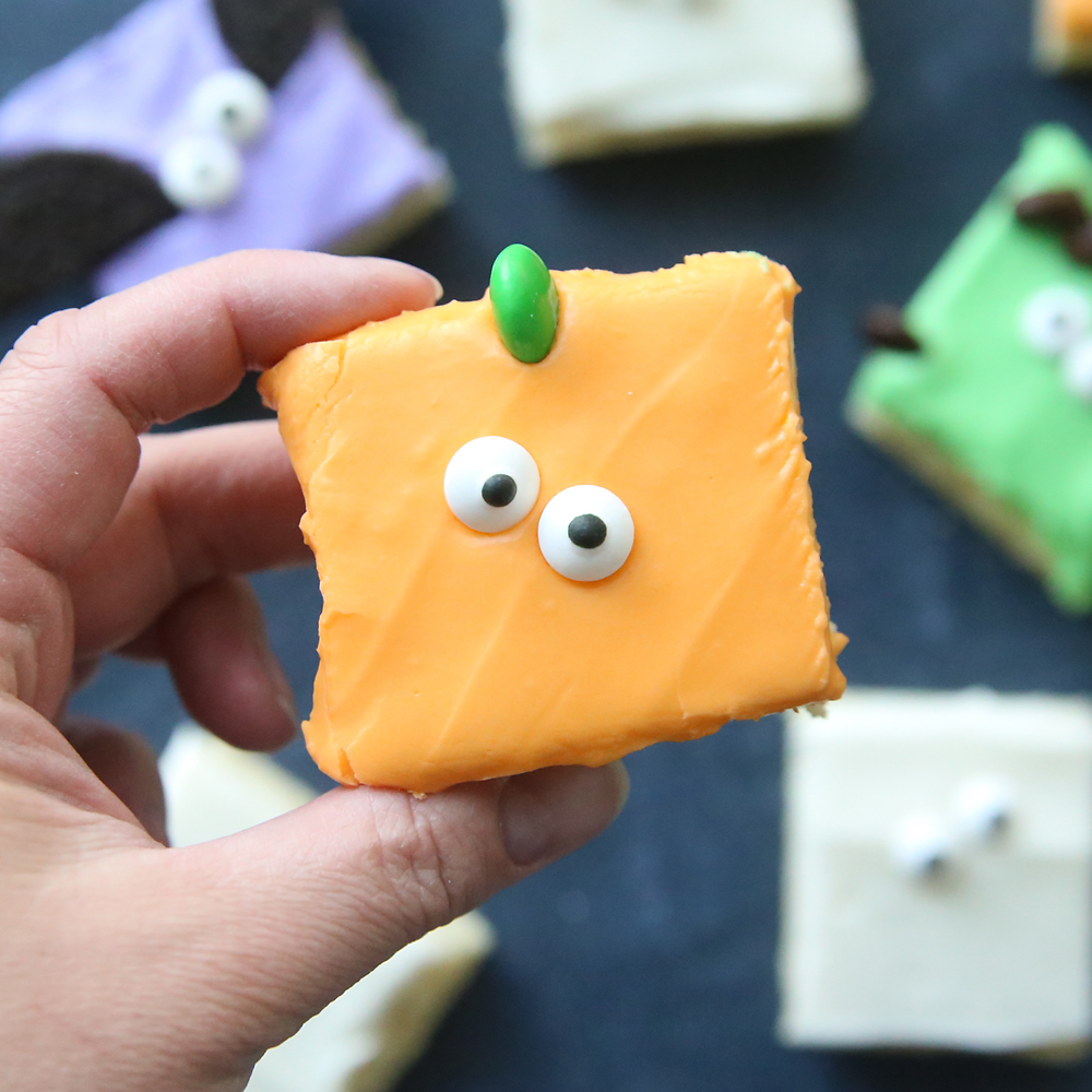 Hand holding a sugar cookie bar with orange frosting, a green M&M on top and two candy eyes