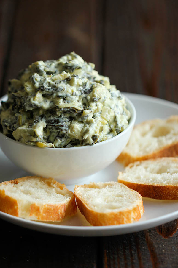 Spinach artichoke dip piled high in a bowl, on a plate with slices of baguette