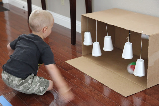 A child rolling a ball toward a cardboard box that has styrofoam cups hanging inside