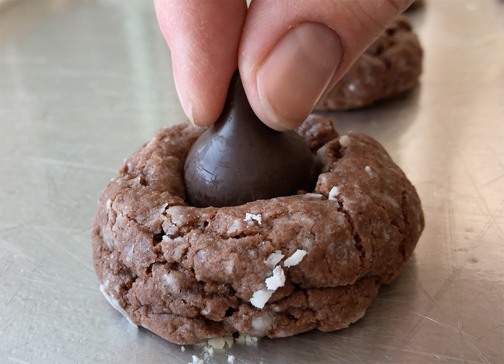 Hand pressing a chocolate mint Hershey kiss onto baked chocolate cookie