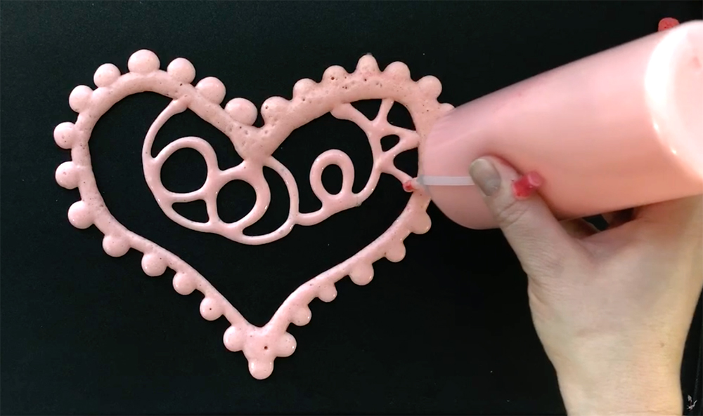 Hand using squeeze bottle to draw with pink pancake batter on a griddle