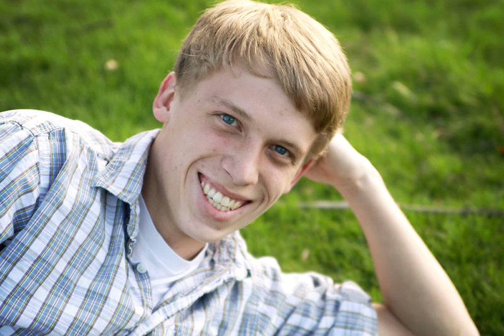 A young man is laying on his side in the grass with his head on his fist