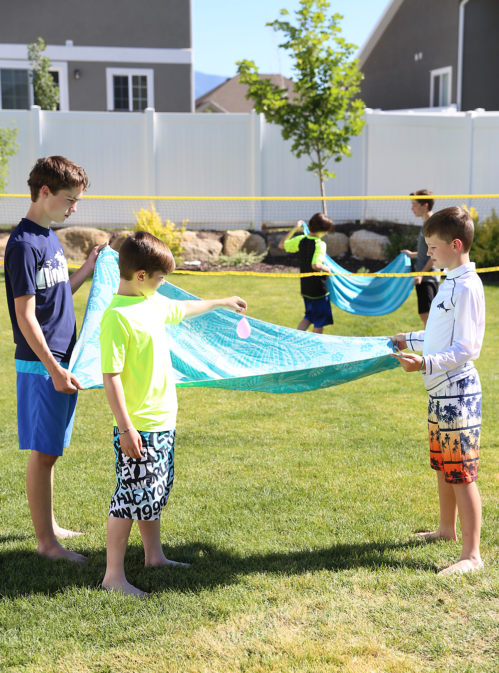 Two boys holding a beach towel while a third boy puts a water balloon on the towel