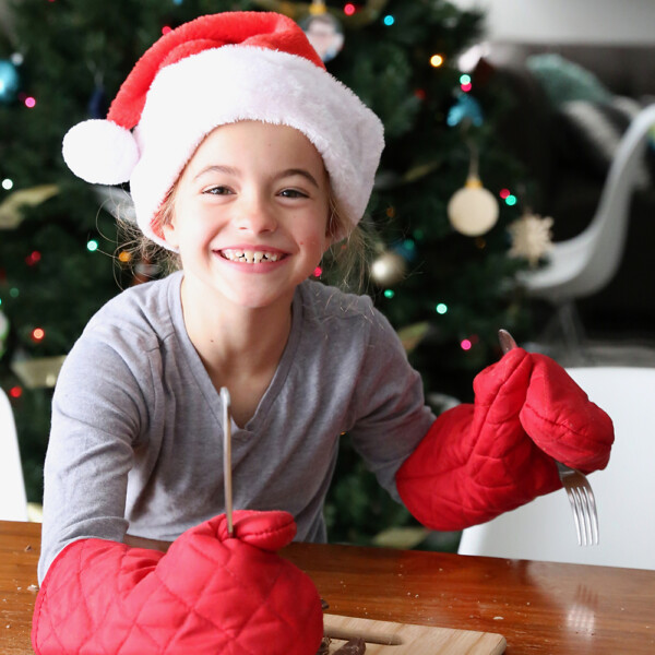 Girl in Santa hat and oven mitts cutting chocolate bar with a fork and knife