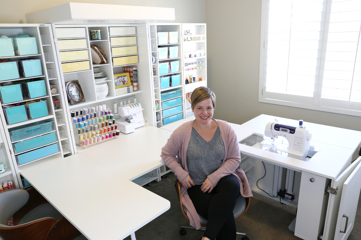 A person sitting at a desk in front of set of craft shelves, with a sewing machine and multiple bins in it