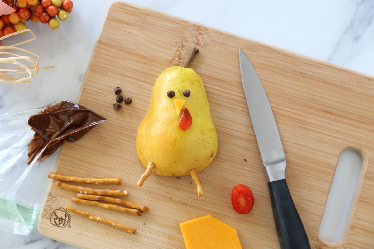 Pear half decorated to look like a turkey with chocolate dots for eyes, small cheese triangle for nose, slice of tomato for waddle and pretzel stick legs