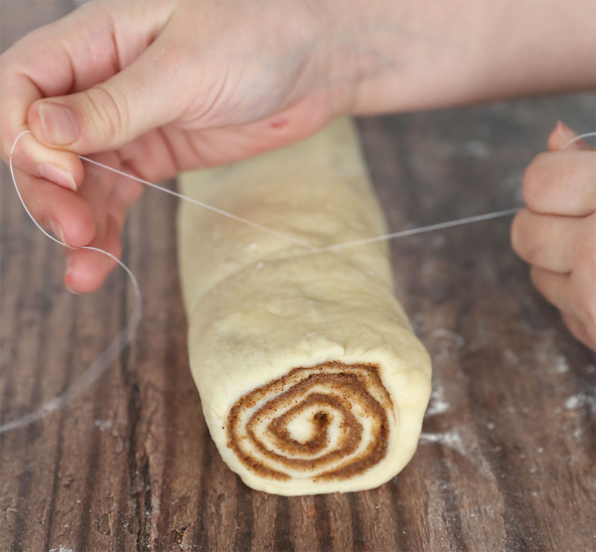 Hands using thread to cut a roll of dough into cinnamon rolls