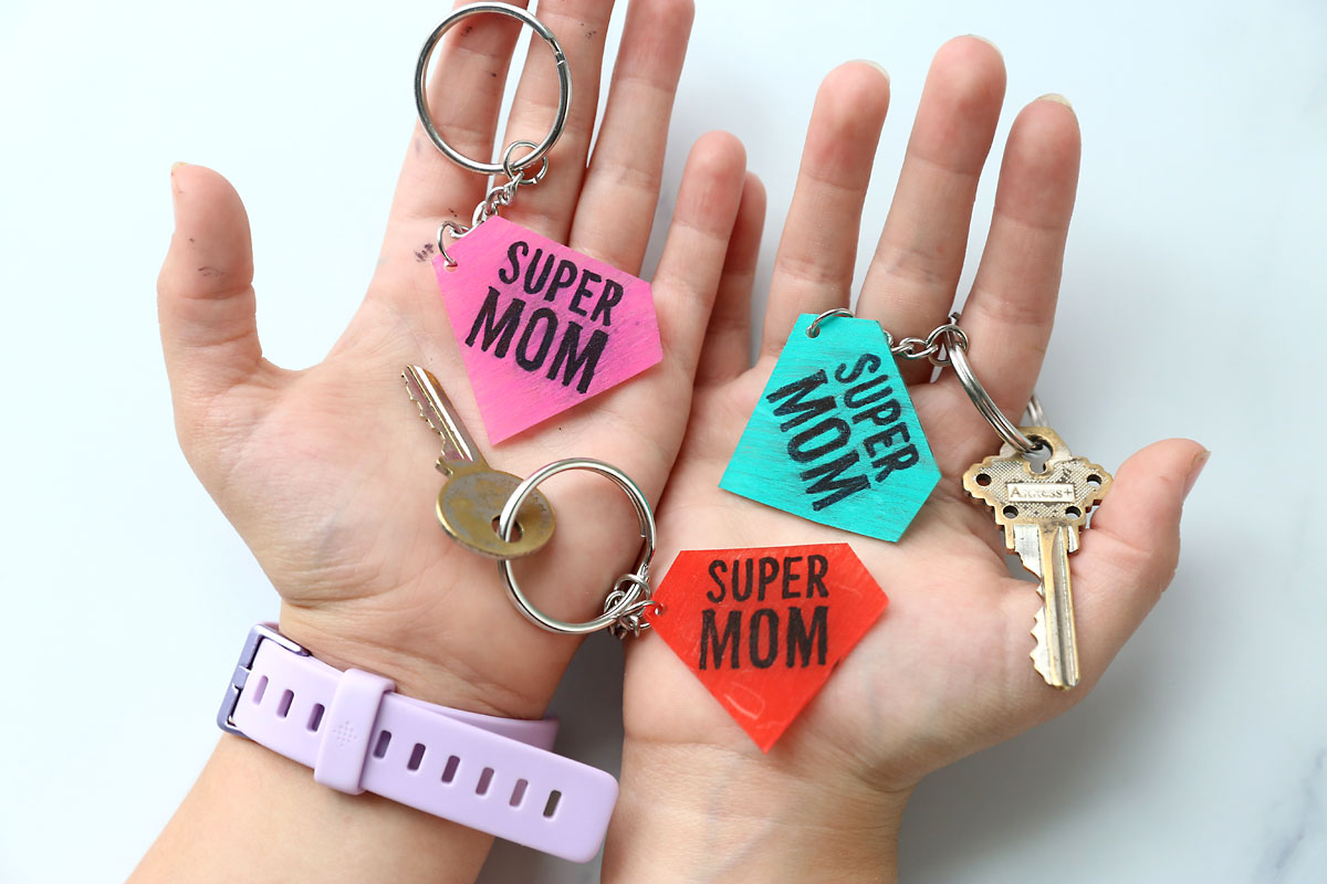 Hands holding DIY shrinky dink keychains that say supermom