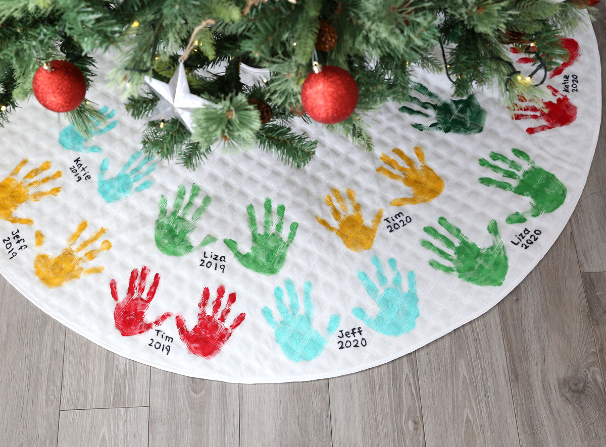 Christmas tree skirt decorated with handprints and names, lying under a Christmas tree