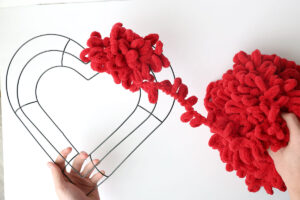 Hand holding a heart shaped wreath form wrapping red loop yarn around it