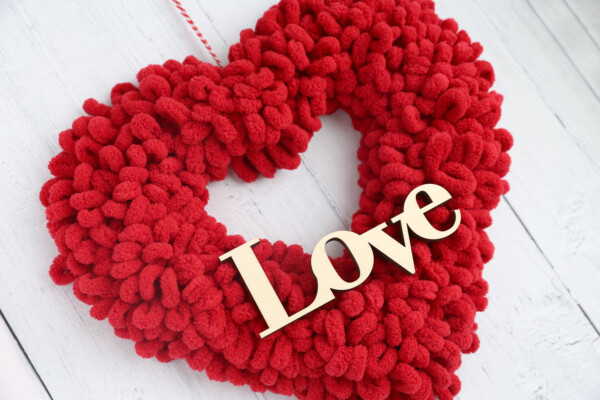 Heart shaped wreath covered in red loop yarn with a wooden cutout of the word Love on it and hanging from twine