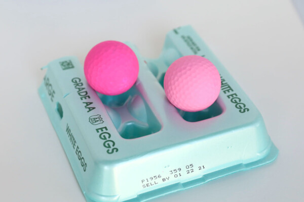 Two golf balls painted pink drying on top of an egg carton