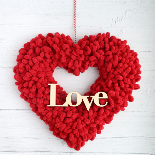 Heart shaped wreath covered in red loop yarn with a wooden cutout of the word Love on it and hanging from twine