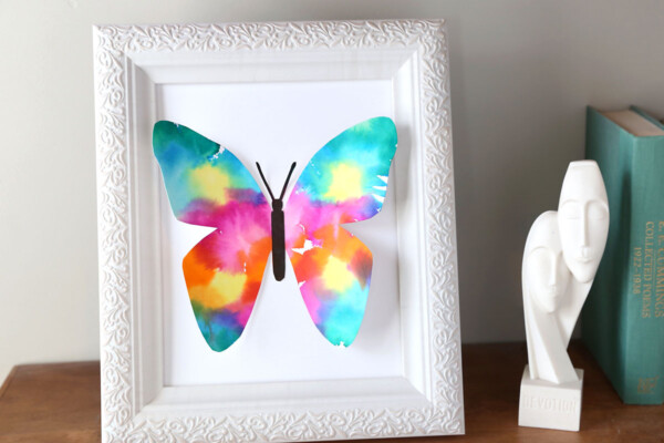 Colorful butterfly made of paper in a white picture frame on a shelf with a small white decorative figurine