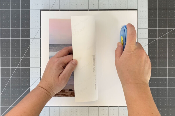 Square of white foam core with a photo centered on it, hand lifting the right half of the photo to put adhesive on the board